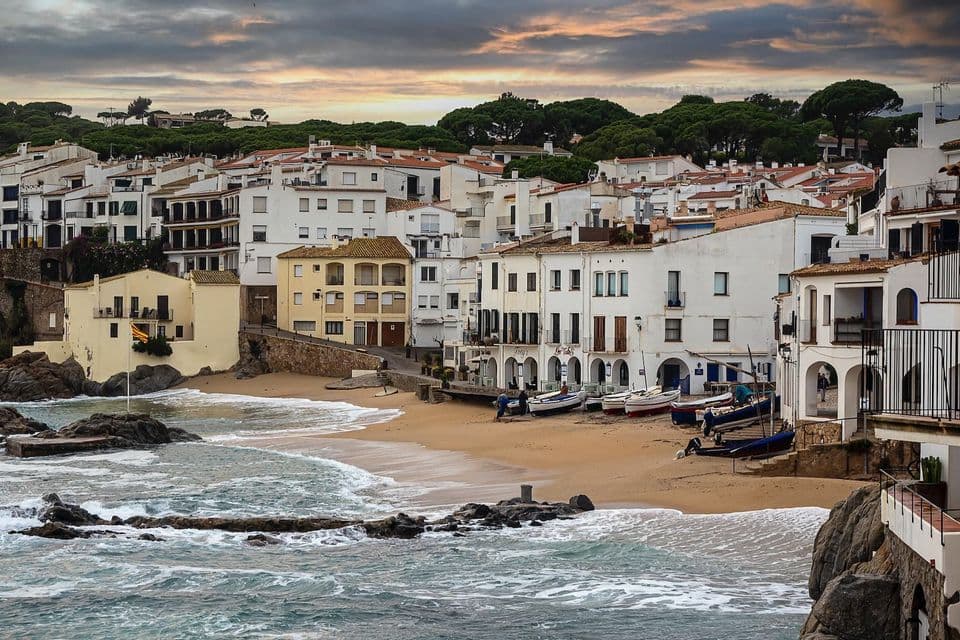 Des bâtiments blancs d'un village côtier bordent une crique de sable avec des bateaux sur le rivage et des vagues se brisant sur les rochers.