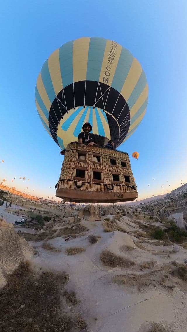 A wide-angle selfie of a person in a hot air balloon flying over a rocky landscape at sunrise, with dozens of other balloons in the sky.