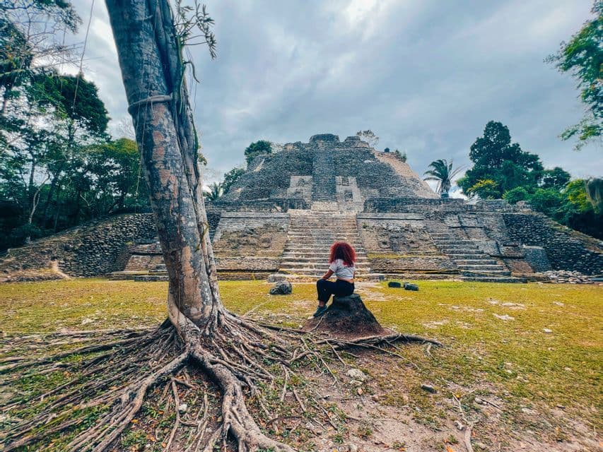 Una persona dai capelli rossi siede su una roccia, osservando una grande piramide di pietra terrazzata, circondata da alberi sotto un cielo nuvoloso.