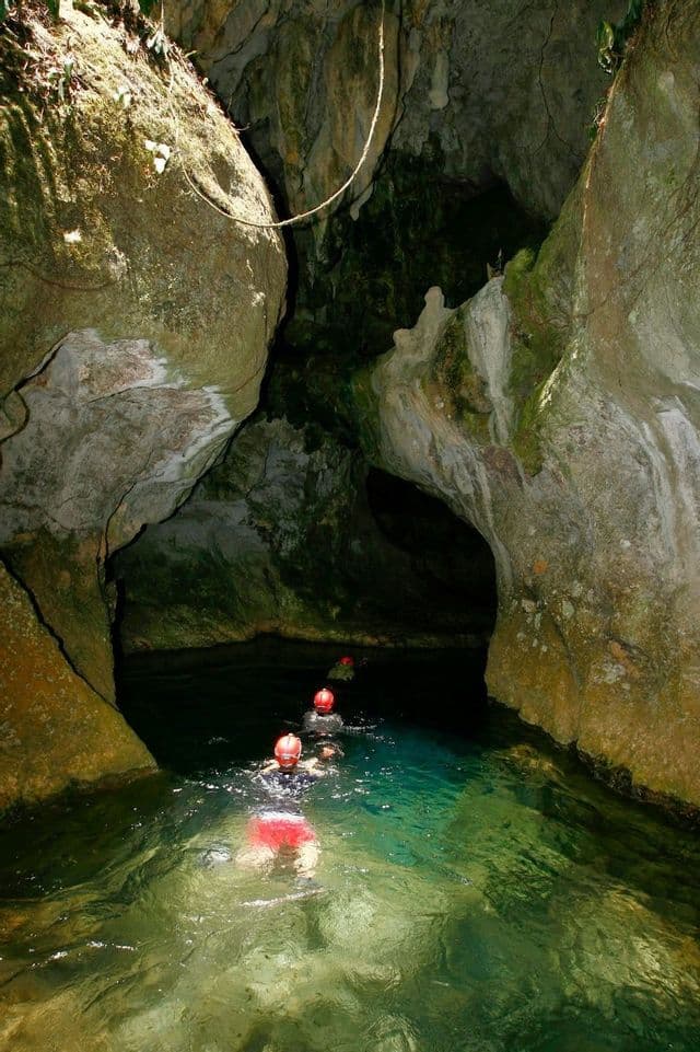 Un viaggio di gruppo WeRoad con caschi rossi nuota in fila indiana nelle acque limpide e turchesi di una grotta illuminata dal sole.