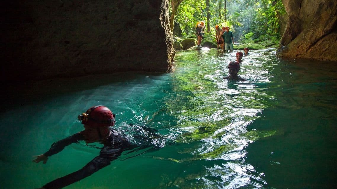 Un gruppo WeRoad in viaggio, con i caschi, nuota e guada in un fiume turchese che esce da una grotta e si addentra nella giungla.