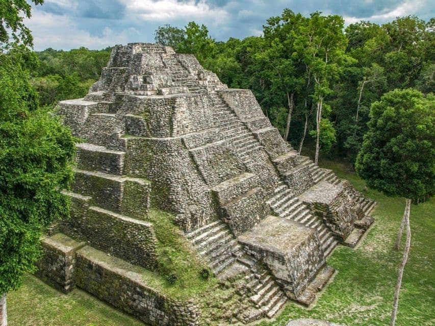 Un'antica piramide in pietra con più livelli e scalinate si erge in una fitta giungla verde sotto un cielo nuvoloso.