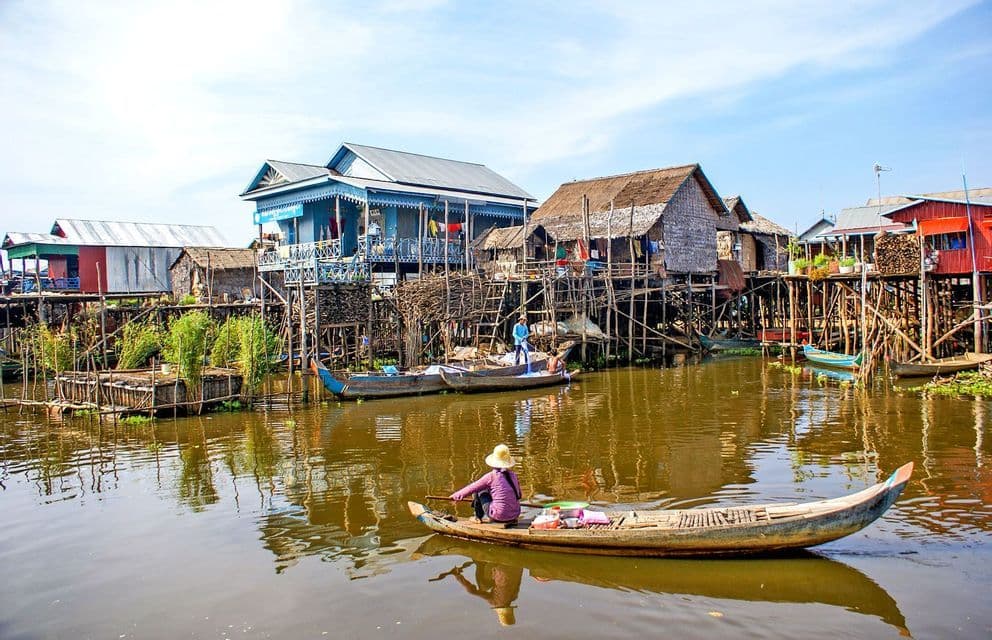 A person in a small boat paddles through a village of colorful houses built on stilts over the water.