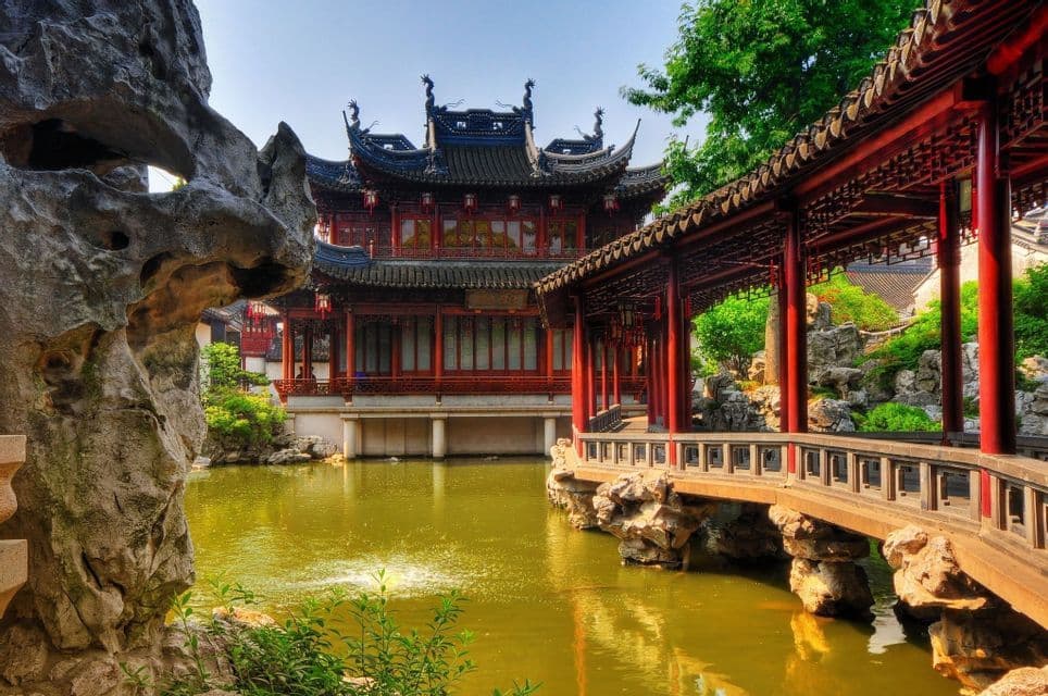 Una tradizionale pagoda cinese si erge su uno stagno accanto a un ponte rosso coperto in un giardino classico con rocce e alberi.