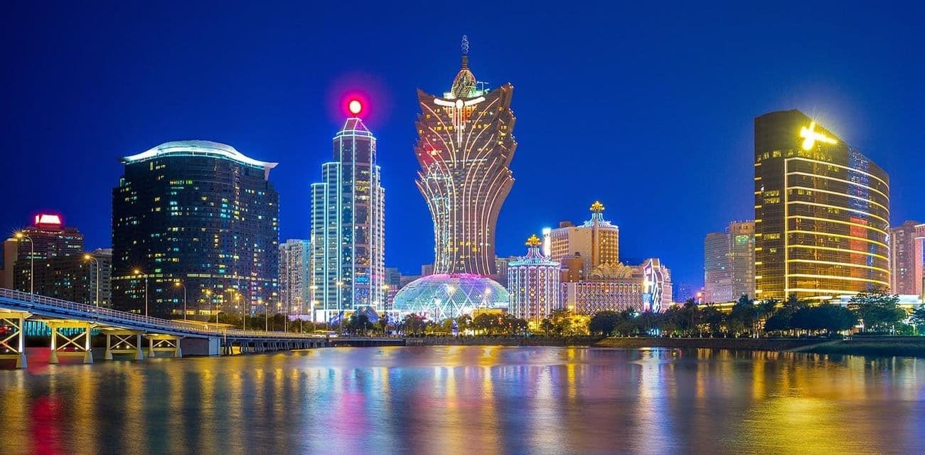 Grattacieli illuminati costeggiano un lungomare di notte, con un ponte sulla sinistra e luci colorate che si riflettono sull'acqua.