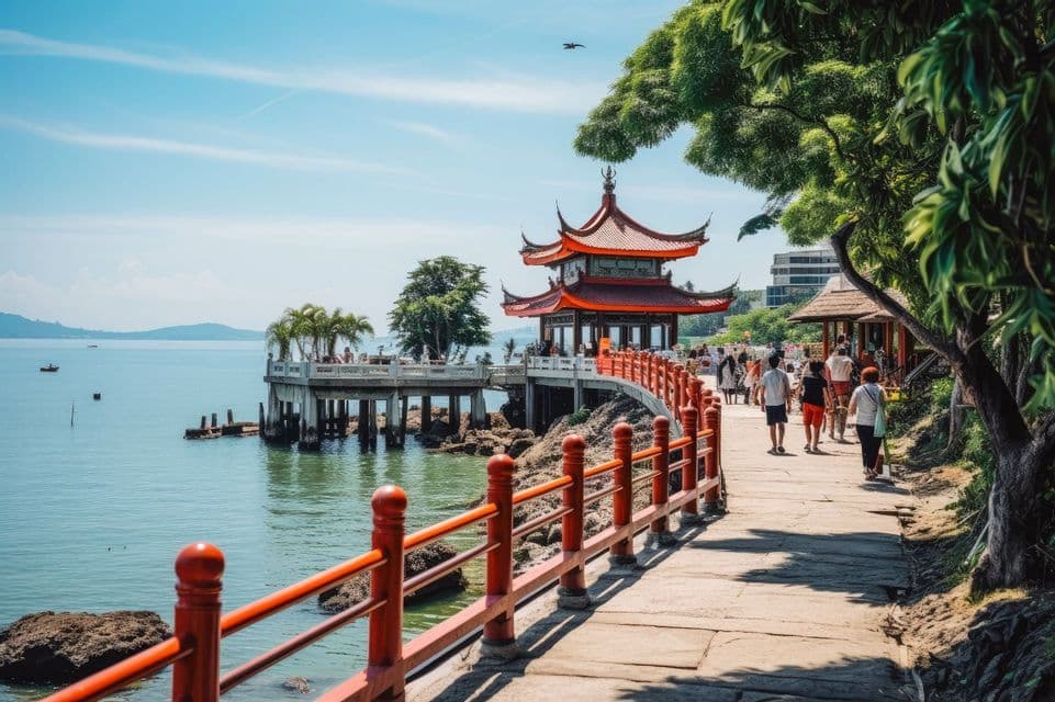 Persone camminano su un sentiero costiero con una ringhiera rossa verso una tradizionale pagoda costruita su un molo sull'acqua.