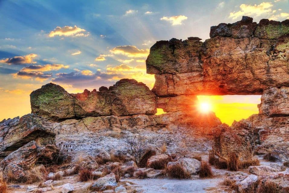 El sol poniente brilla a través de una ventana natural en una gran formación rocosa, con rayos de sol que se irradian hacia el cielo nublado.