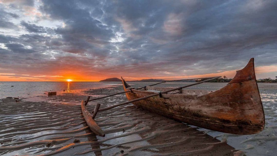 Una canoa de madera con balancín descansa en una playa de arena ondulada durante la marea baja y un atardecer nublado.