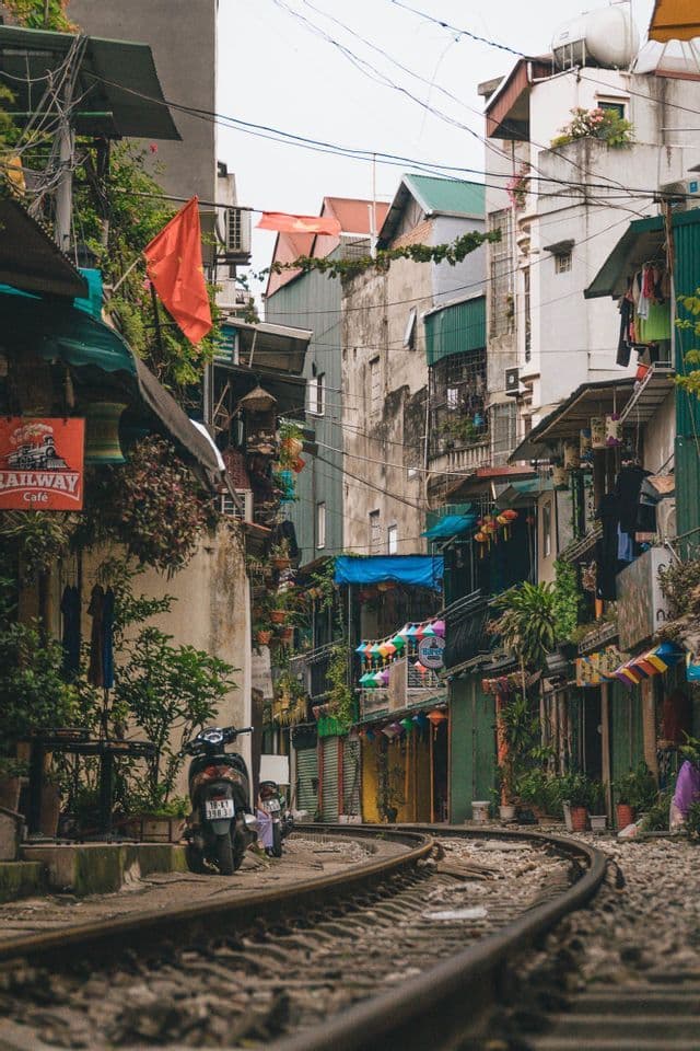 Un'inquadratura dal basso di binari ferroviari che si snodano in una strada stretta, fiancheggiata da edifici, negozi e uno scooter parcheggiato.