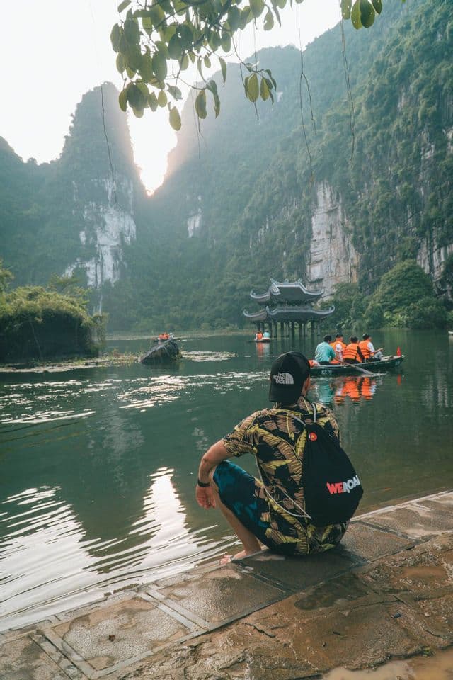 Un uomo con uno zaino WeRoad si accovaccia ai margini di un fiume, osservando un gruppo in barca vicino a una pagoda e alte montagne carsiche.