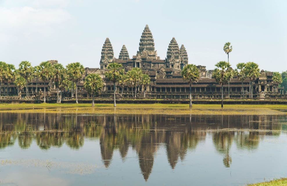 Un grande tempio antico con diverse torri si riflette in uno specchio d'acqua calmo, dietro una fila di palme verdi.