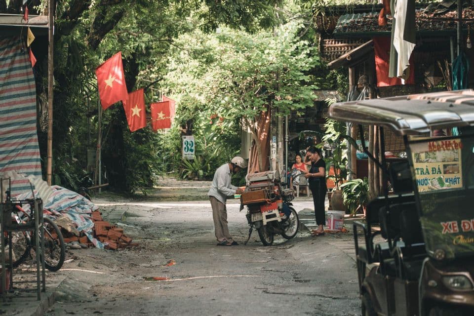 Un uomo sistema uno scooter carico su una strada rustica alberata, con bandiere rosse con stelle gialle sullo sfondo.