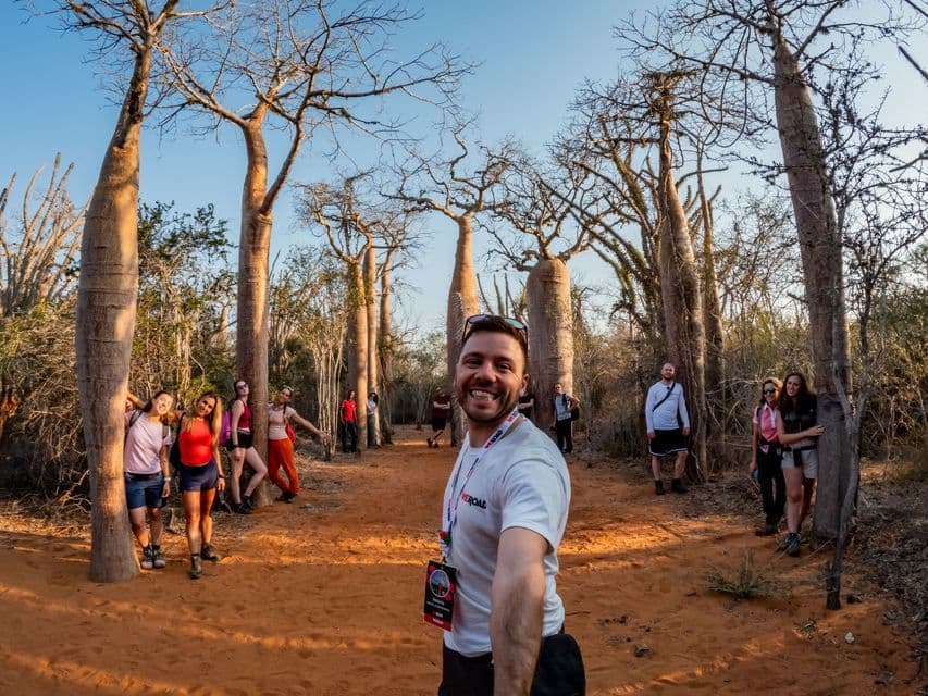 Un uomo si scatta un selfie con il suo gruppo di viaggio WeRoad mentre posano tra grandi alberi di baobab su un sentiero di terra rossa.