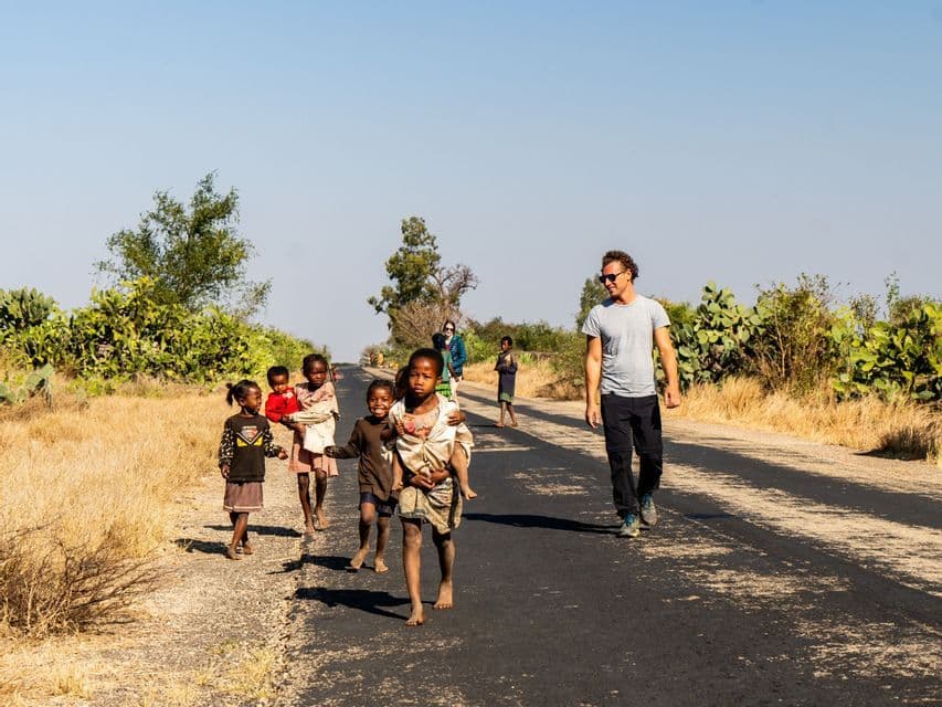 Un uomo di un viaggio di gruppo WeRoad cammina con diversi bambini locali a piedi nudi lungo una strada soleggiata e asfaltata in campagna.