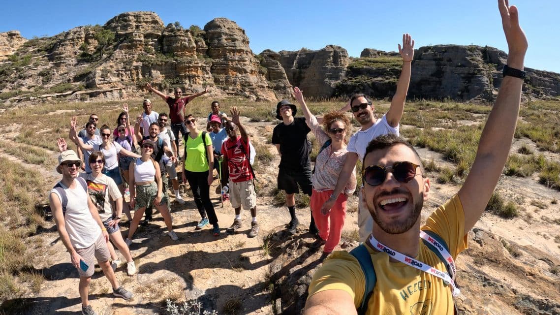 Un uomo si scatta un selfie del suo viaggio di gruppo WeRoad mentre fanno escursioni e salutano in un paesaggio roccioso sotto un cielo sereno.