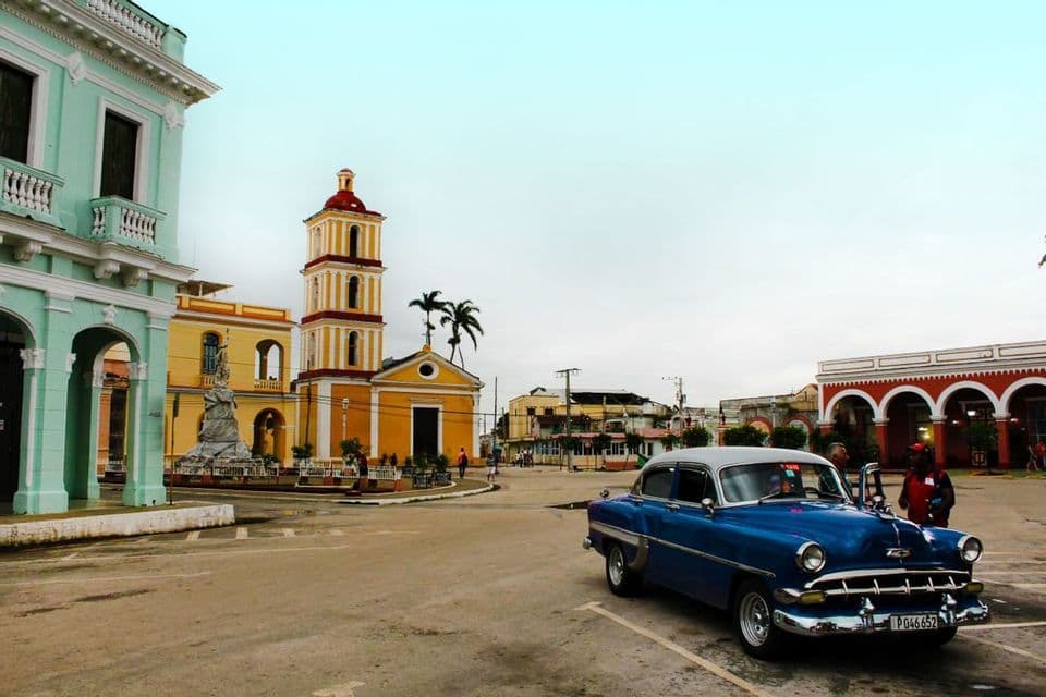Une voiture bleue classique garée sur une place de ville, avec des bâtiments coloniaux colorés et une église jaune en arrière-plan.