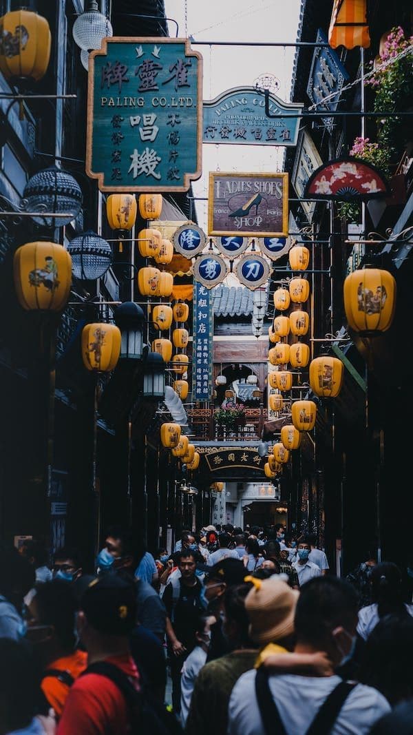 Una grande folla di persone passeggia lungo uno stretto vicolo cittadino, illuminato da lanterne gialle appese e insegne di negozi tradizionali.