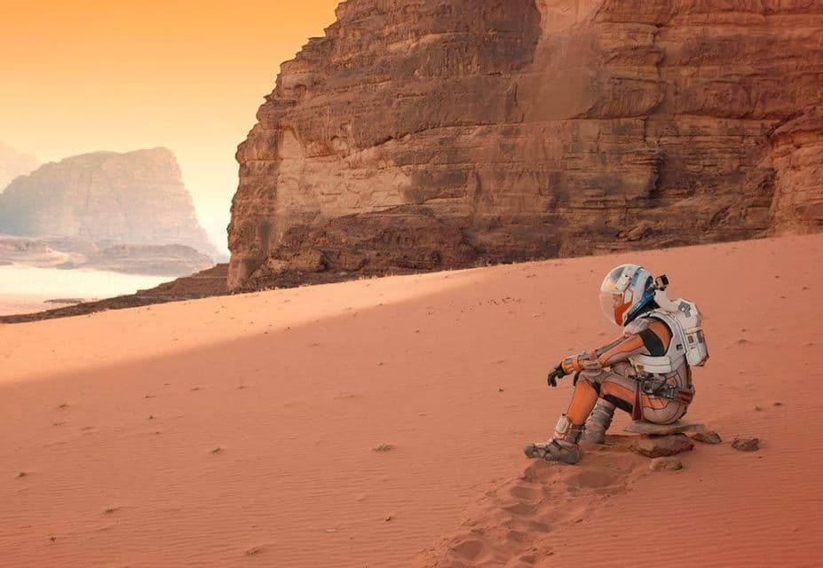 Un astronauta in tuta spaziale completa siede su rocce su una duna di sabbia rossa, con grandi formazioni rocciose sullo sfondo.