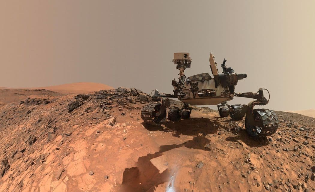 Il rover Curiosity della NASA è posizionato su un terreno roccioso e rosso su Marte, con colline desolate visibili sotto un cielo polveroso.