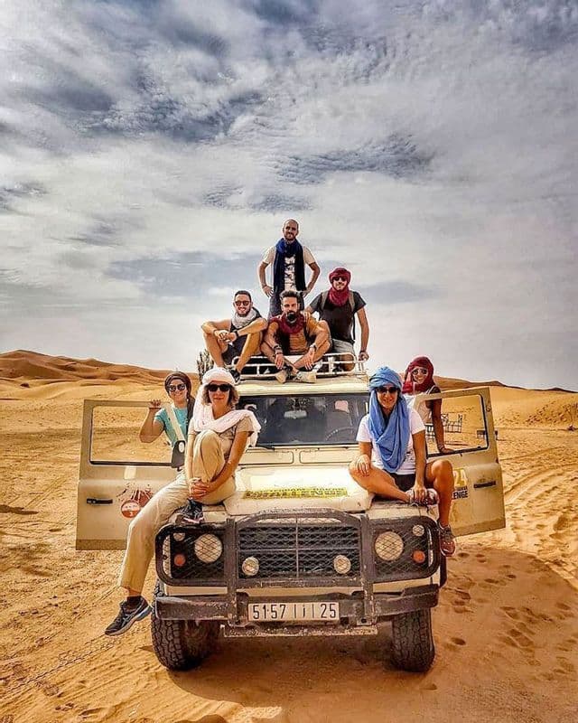 Un viaje en grupo de WeRoad posa sobre y alrededor de un vehículo todoterreno blanco en un desierto arenoso bajo un cielo nublado.