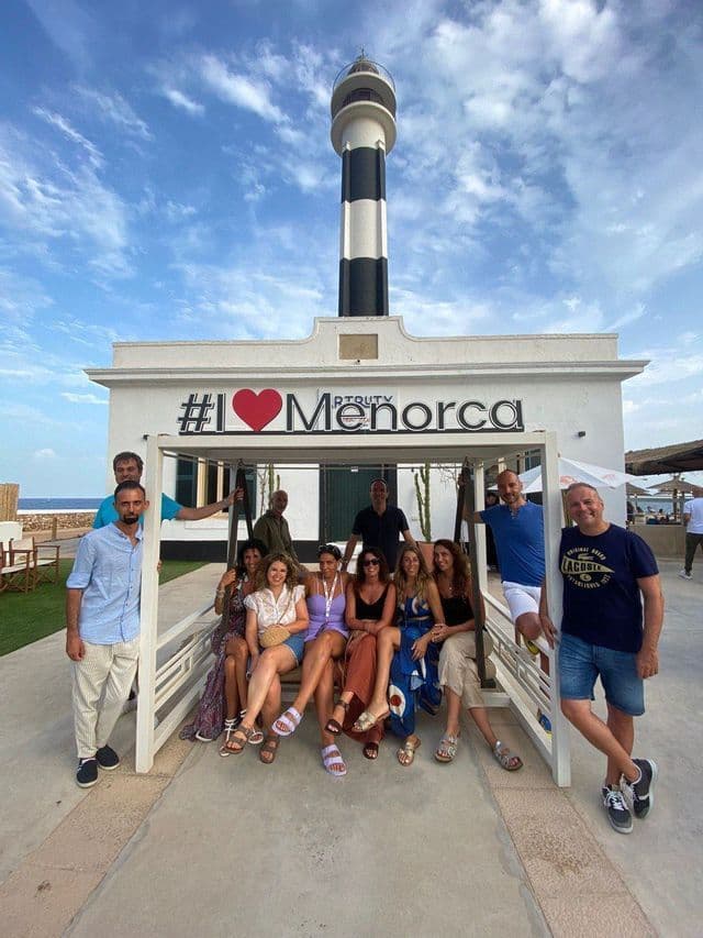 Eine WeRoad-Gruppe posiert auf einer großen weißen Bank vor einem Schild mit der Aufschrift „#I ❤️ Menorca“, dahinter ein gestreifter Leuchtturm.