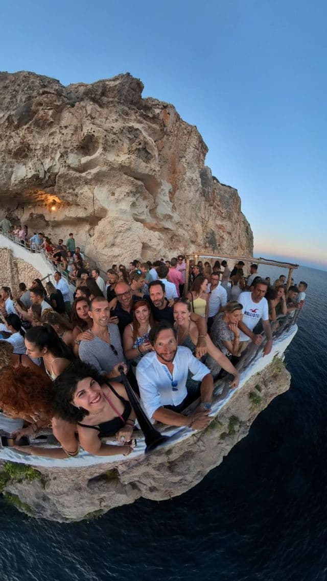 Eine WeRoad Gruppenreise macht ein Weitwinkel-Selfie auf einer belebten Bartrasse, die in einen Felsen über dem Meer gebaut ist.
