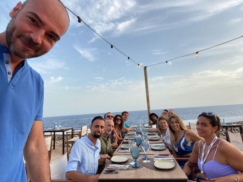 Eine WeRoad-Gruppe macht lächelnd ein Selfie an einem langen Esstisch in einem Außenrestaurant am Meer.