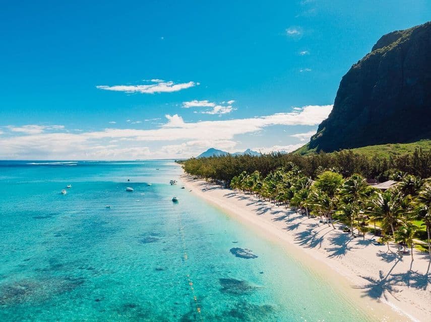 Vista aerea di una costa tropicale con spiaggia di sabbia bianca, acqua turchese e una lussureggiante montagna verde sotto un cielo azzurro chiaro.