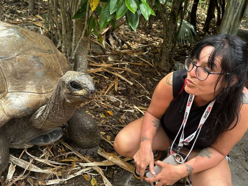 Una donna con occhiali e un laccetto WeRoad si accovaccia faccia a faccia con una tartaruga gigante in una foresta.