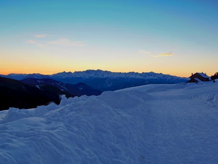 A cabin sits on a snowy mountainside at sunset, with a distant snow-capped mountain range against a clear gradient sky.