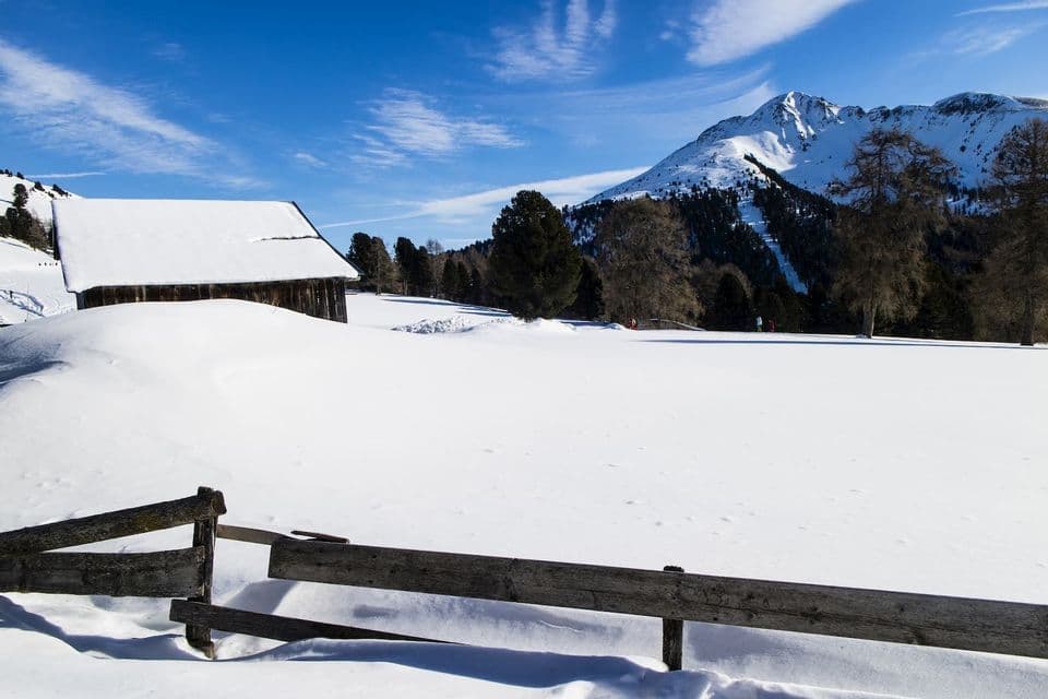Eine rustikale Holzhütte und ein Zaun sind in einer tief verschneiten Berglandschaft unter strahlend blauem Himmel.