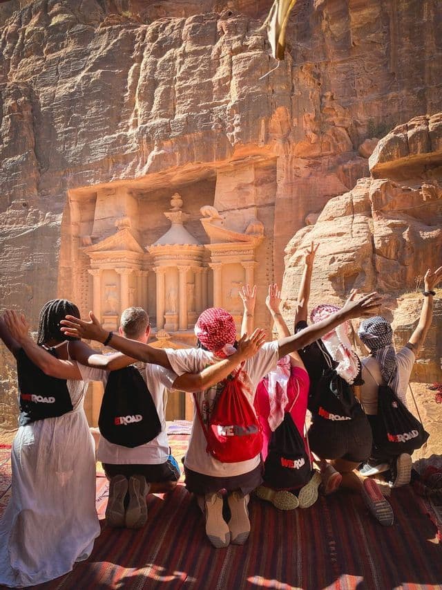 Eine WeRoad-Gruppe kniet auf einem Teppich mit erhobenen Armen und blickt auf einen großen, in einen Sandsteinfelsen gehauenen Tempel.
