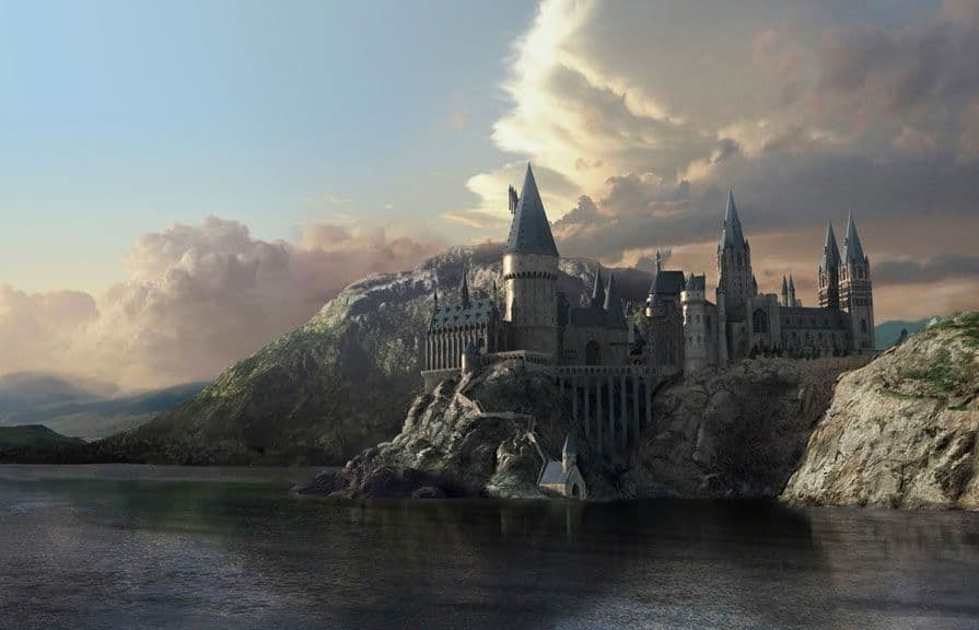 Un gran castillo con muchas agujas se alza sobre un acantilado rocoso que domina un lago, con montañas visibles al fondo bajo un cielo nublado.