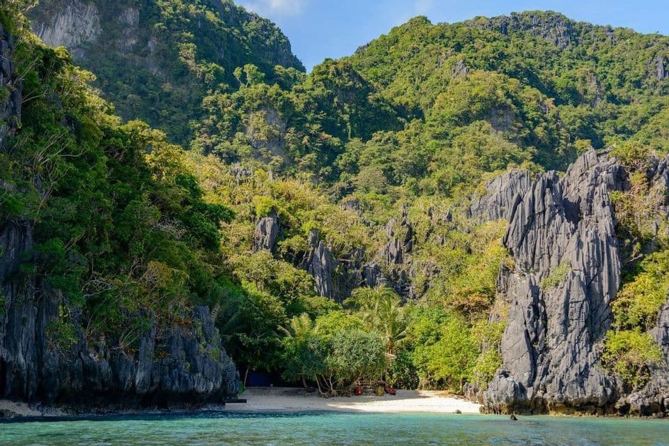 Una caletta sabbiosa appartata con acqua turchese, circondata da ripide formazioni rocciose ricoperte dalla giungla sotto un cielo azzurro.