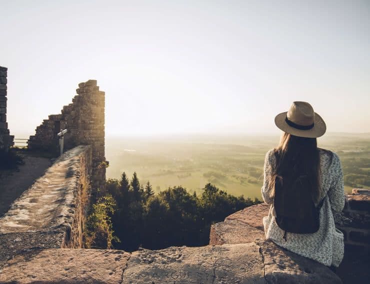 Una donna con cappello e zaino siede sul bordo di una rovina di pietra, osservando una valle e una foresta illuminate dal sole.