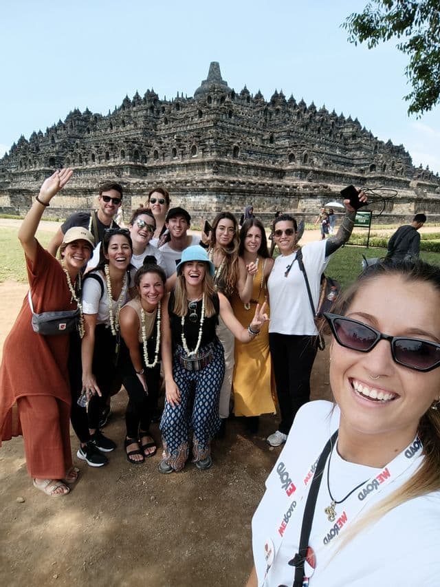 Un grupo de WeRoad sonríe y posa para una selfie frente a un gran templo de piedra antiguo en un día soleado.