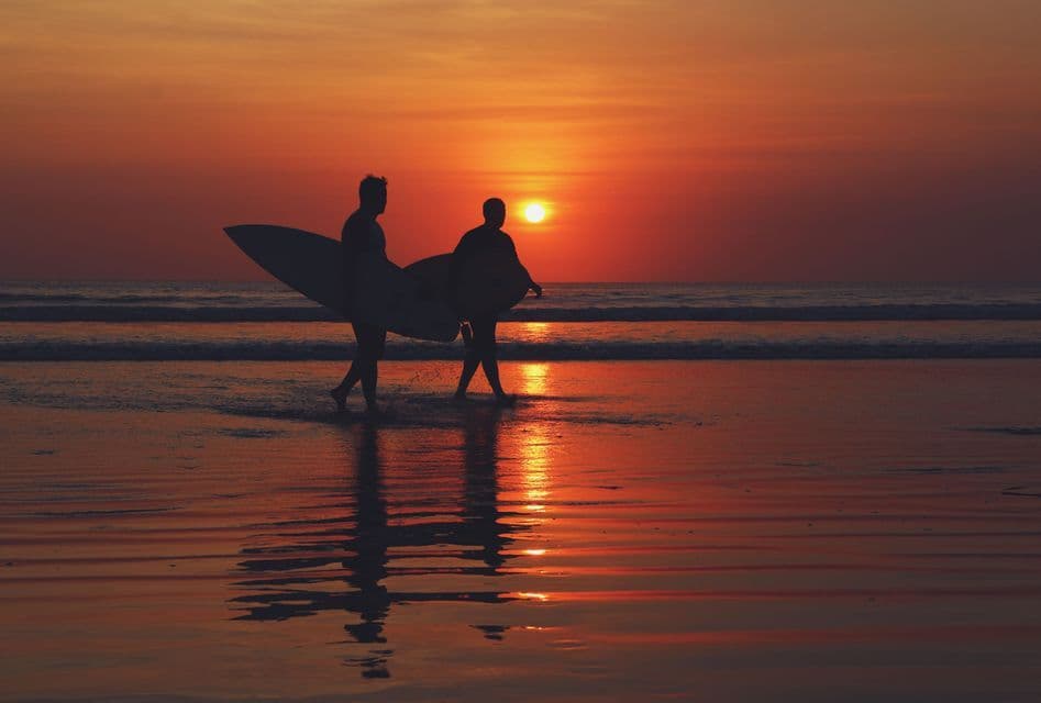 Dos surfistas silueteados por el sol poniente llevan sus tablas de surf mientras caminan por la orilla.