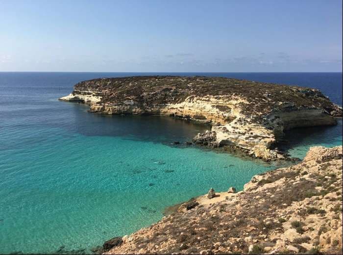 Un isolotto roccioso e coperto di macchia mediterranea si trova in una baia con acqua bassa e turchese che contrasta con il blu profondo del mare.