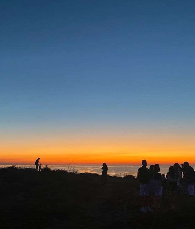 Sagome di un viaggio di gruppo WeRoad su una collina che osservano il tramonto arancione e blu sul mare.