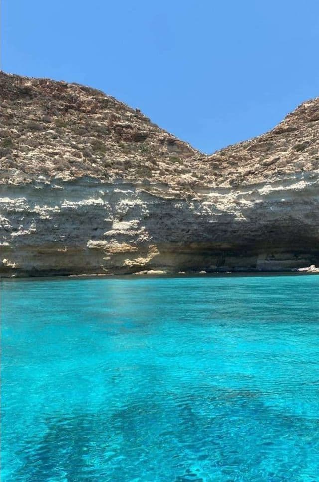 Acque turchesi cristalline lambiscono una scogliera rocciosa stratificata, con una grotta marina alla base, sotto un cielo azzurro brillante.