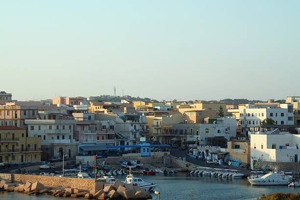 Una città costiera baciata dal sole, con edifici fittamente raggruppati, si erge sopra un piccolo porto pieno di barche ormeggiate.
