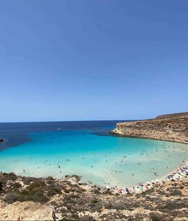 Una cala affollata vista dall'alto, con persone che nuotano in acque limpide e turchesi e scogliere rocciose.