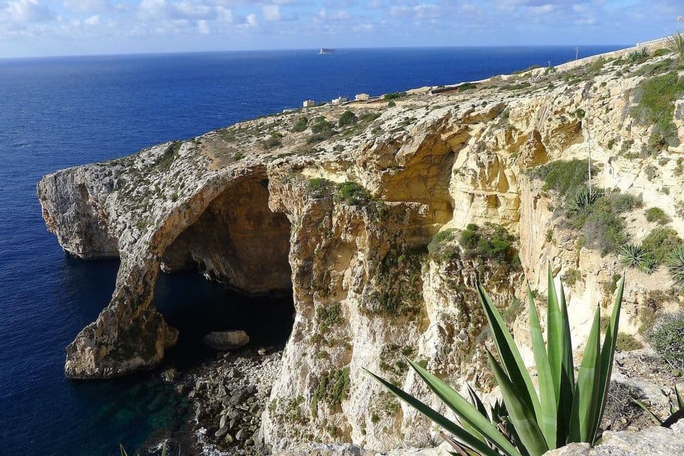 Veduta di un grande arco naturale sul mare formato da scogliere calcaree lungo una costa con acqua di un blu intenso.