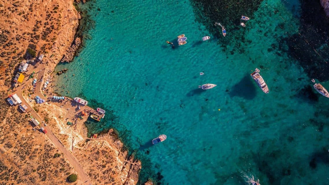 Una vista aerea dall'alto di una cala rocciosa con acqua turchese cristallina, piena di barche e persone su una piccola spiaggia.