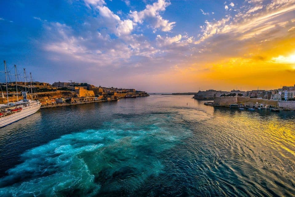 Una vista dall'acqua di un porto storico della città al tramonto, con una grande nave ormeggiata e una scia turbolenta in primo piano.