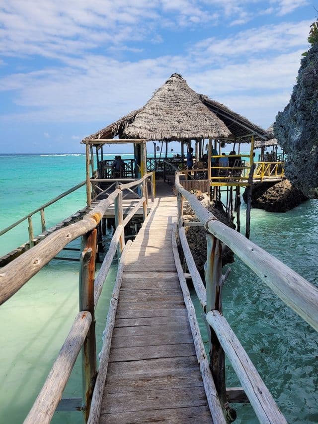 A wooden pier leads to a thatched-roof restaurant built on stilts over turquoise water under a partly cloudy sky.