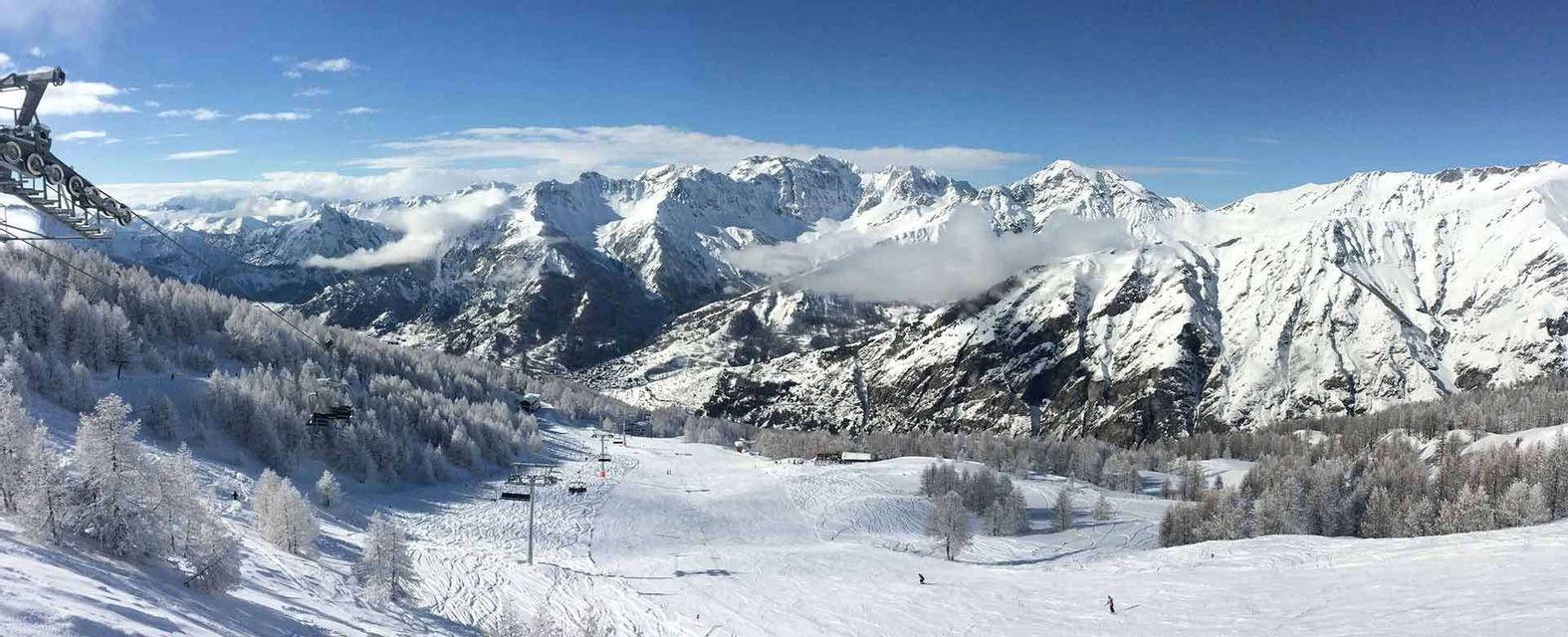 Una vista panoramica di un paesaggio montano innevato con un centro sciistico, seggiovie, piste da sci e alberi ghiacciati sotto un cielo blu.