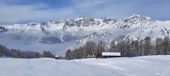 Una baita di legno si trova in un paesaggio innevato di fronte a una vasta catena montuosa innevata sotto un cielo blu.