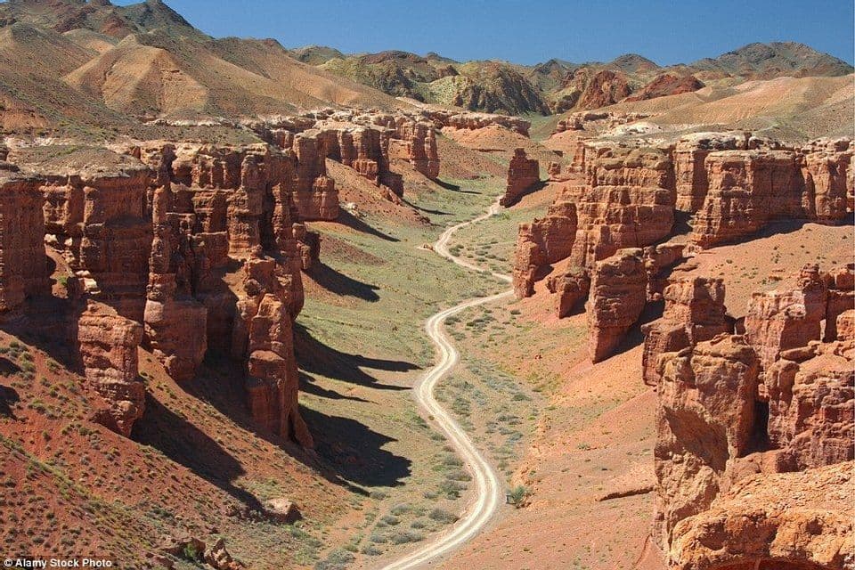 A high-angle view of a winding dirt road running through a deep canyon with red rock formations and green vegetation.