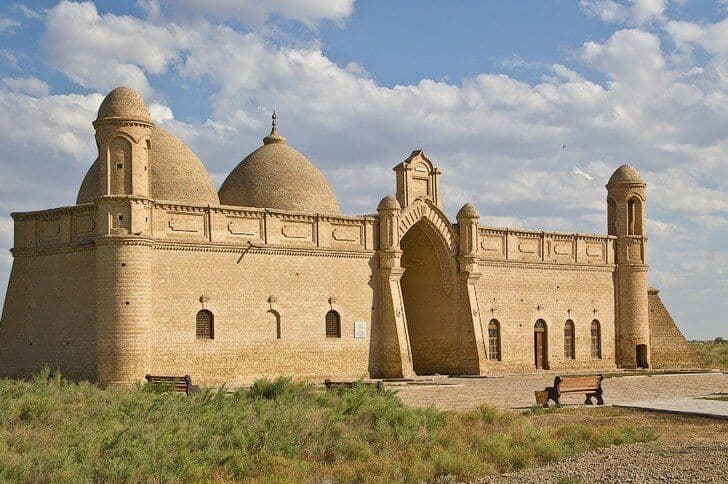 A historical sand-colored building with multiple domes and a large arched entrance sits in a grassy landscape under a cloudy sky.